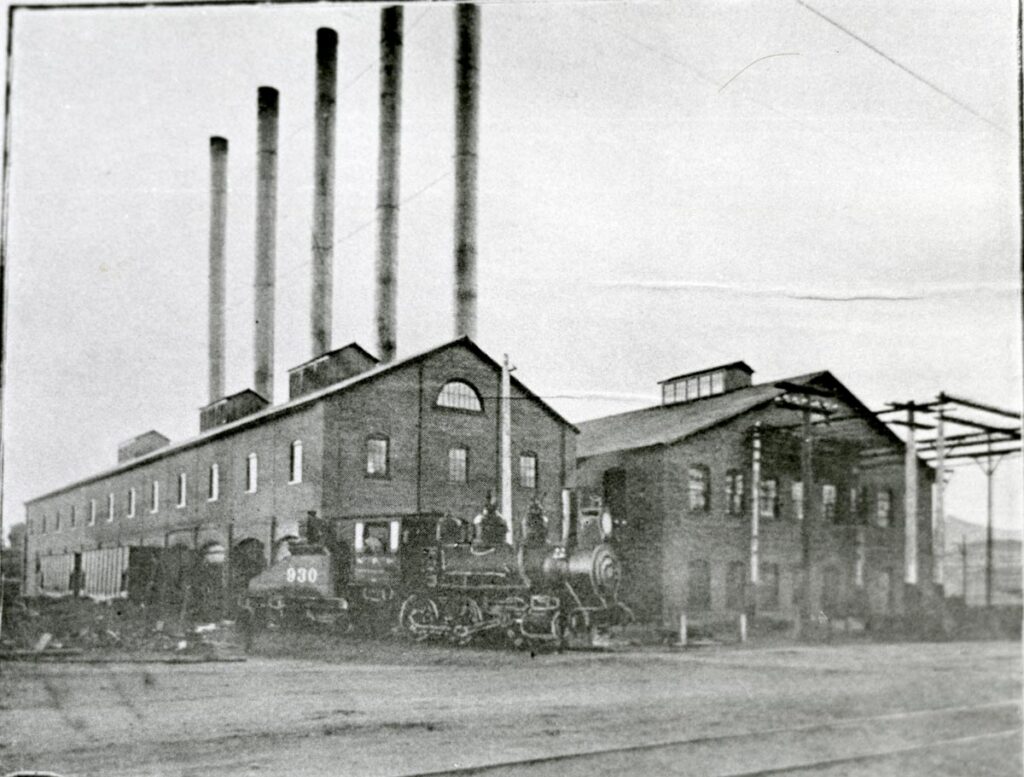 Locomotive of the B. A. & P. Railroad (Butte, Anaconda & Pacific Railroad) is stopped at the side of a plant which supplied electricity for the mines in Butte, MT. South Montana St. Circa 1910